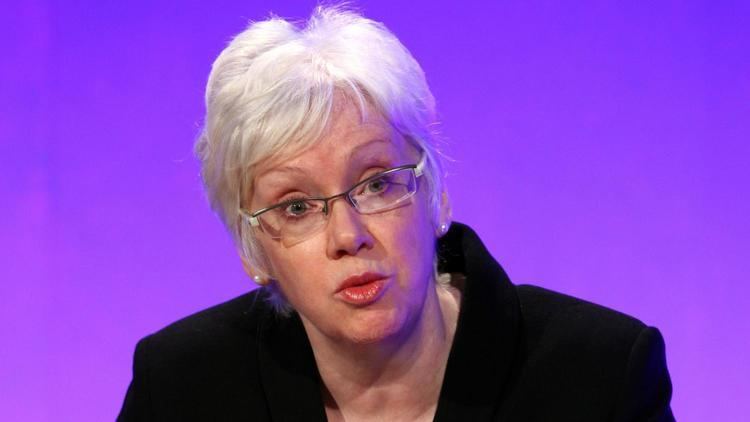 In a purple background, Margaret Ford, Baroness Ford is serious, eyebrows up, has white hair, wearing eyeglasses, pearl earrings and black coat.