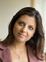 Margarita Simonyan with a tight-lipped smile while looking at something, wearing a cross necklace and a purple top.