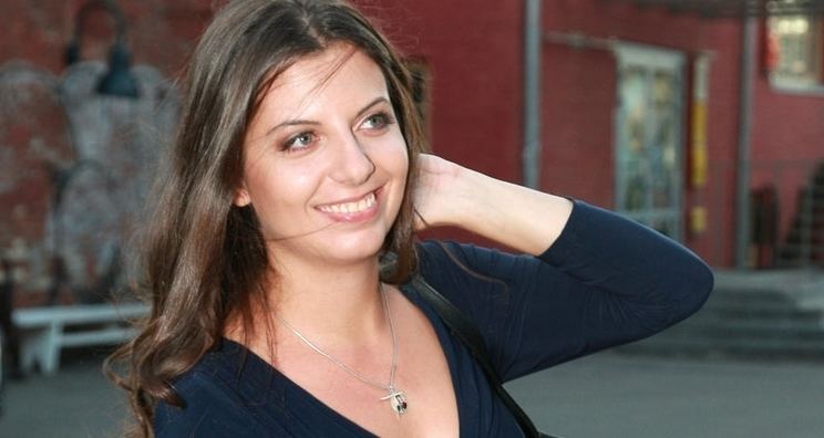Margarita Simonyan is smiling while looking at something and holding her hair, wearing a necklace and a black long sleeve top.
