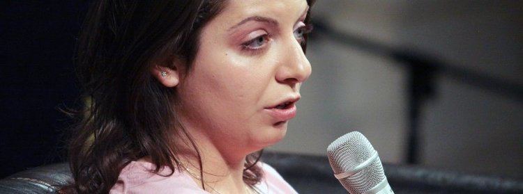 Margarita Simonyan with a serious face while talking in the microphone, wearing earrings, a necklace, and a pink blouse.