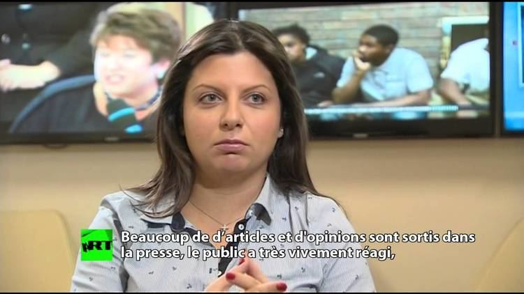 Margarita Simonyan with a serious face while looking at something with televisions in the background. Margarita is wearing a necklace and a gray long sleeve top.