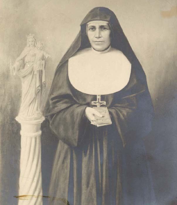 Maria Domenica Mazzarello is smiling while holding a small book with a statue on her left, she is wearing a white head cover under a black nun veil, a long cross necklace under a religious nun habit clothing