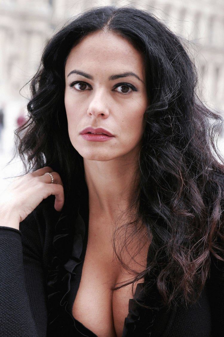 Maria Grazia Cucinotta has a serious-looking face with her long black wavy hair down with a silver ring finger on her right hand, and an exposed cleavage wearing a black ruffled-neck dress.