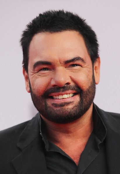 Marian Gold smiling, with a beard and mustache and wearing a black suit and black shirt.