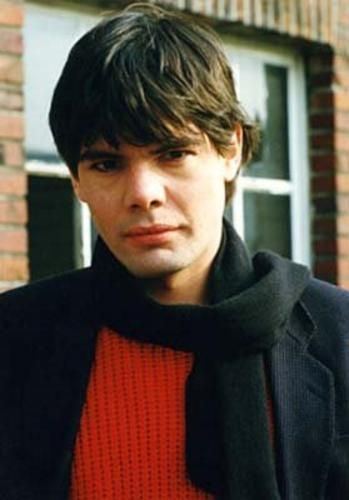Marian Gold with a serious face, wearing a black scarf, black coat, and red shirt.