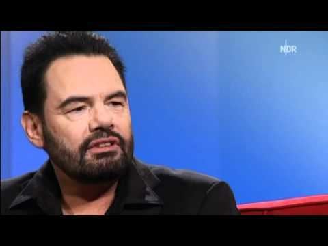 Marian Gold with a beard and mustache and wearing a black polo shirt.