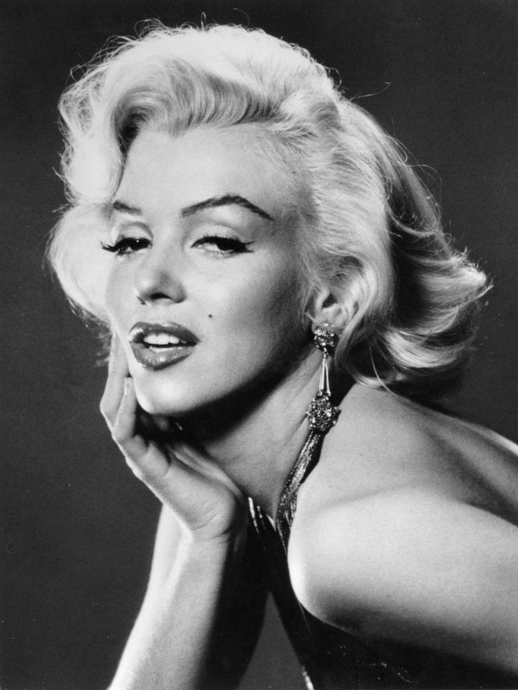 Marilyn Monroe is serious, mouth half opened, has white hair, a mole on her left cheek, right hand on her face, and wears a necklace and a backless dress.