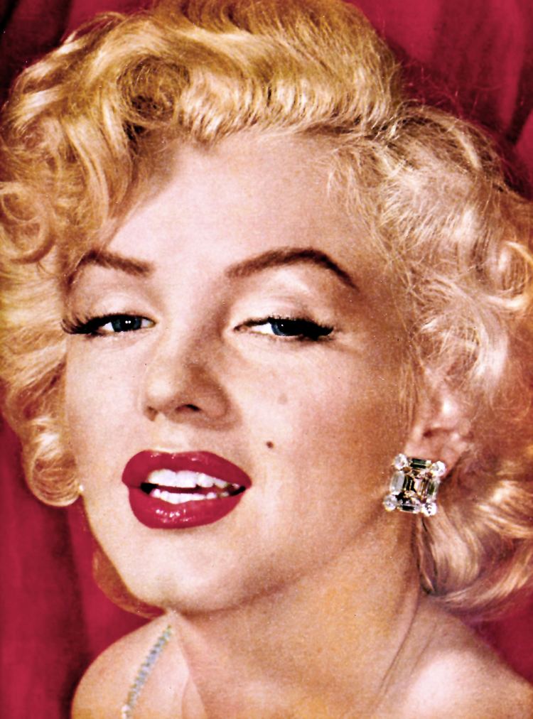 Marilyn Monroe is serious, mouth half opened, has white hair, a mole on her left cheek, and wears white-black earrings.