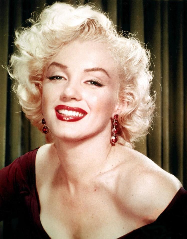 Marilyn Monroe is smiling, has white hair, and a mole on her chest, and wears a red necklace and a red cleavage showing dress.