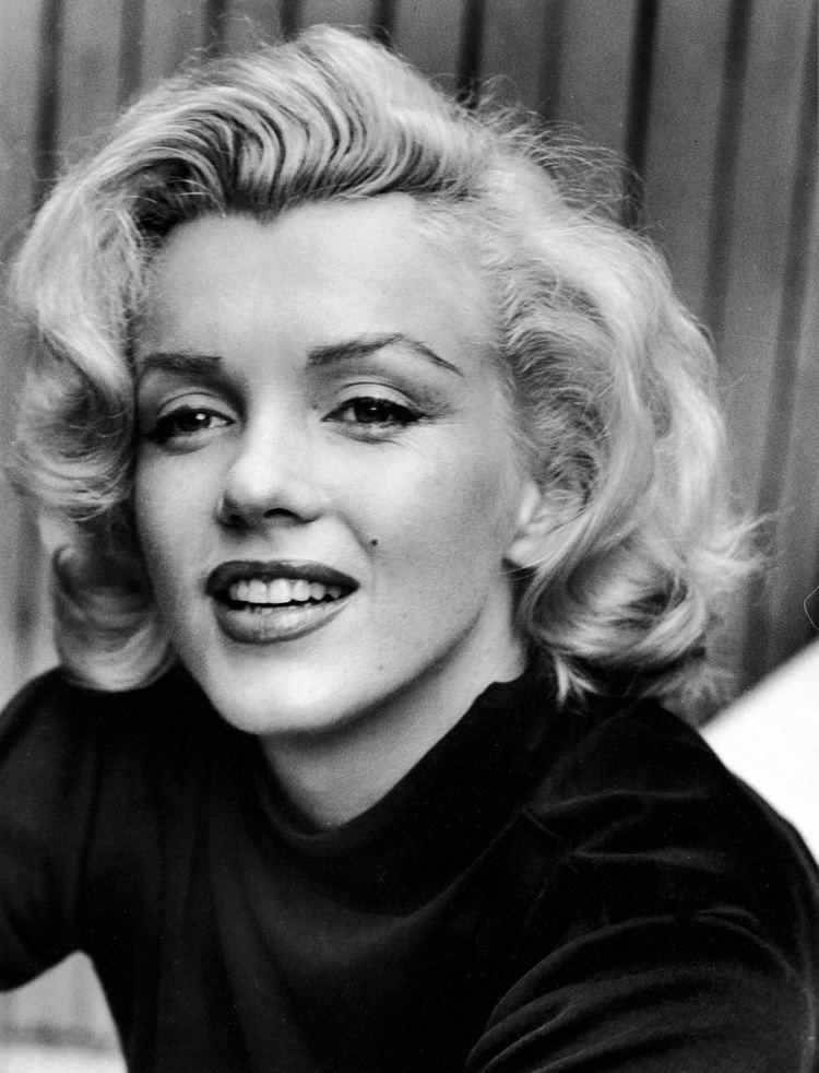 Marilyn Monroe is smiling, has white hair, a mole on her left cheek, and wears a black top.