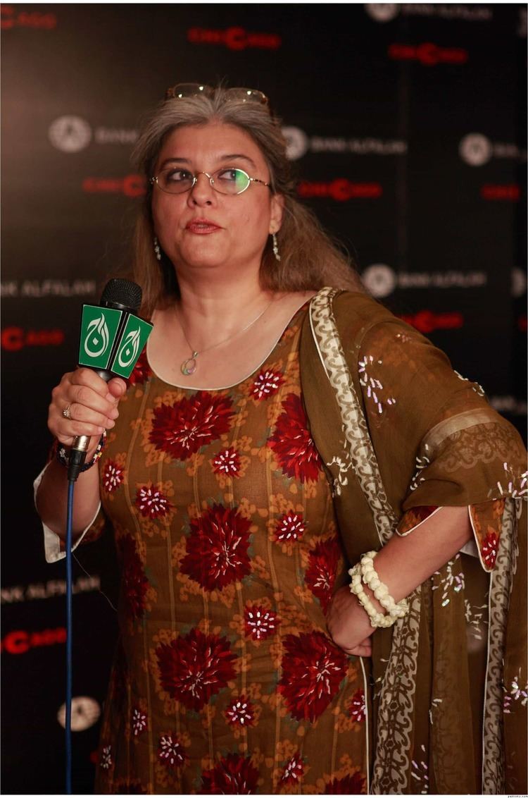 Marina Khan holding a microphone while wearing a brown and red dress, eyeglasses, and some pieces of jewelry