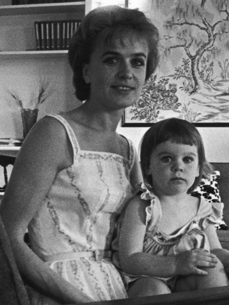 Marina Oswald smiling while carrying her daughter June Lee Oswald. Marina with short hair and wearing a white sleeveless dress while her daughter also wearing a sleeveless dress.