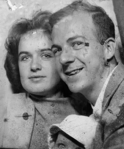 Marina Oswald and Lee Harvey Oswald are smiling while carrying a doll. Marina with short wavy hair and wearing a coat over a knitted turtleneck top while Lee wearing a coat over white long sleeves.