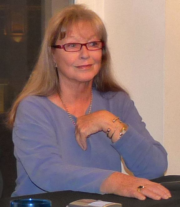Marina Vlady with a tight-lipped smile while wearing a blue long sleeve blouse, eyeglasses, necklace, bracelet, and ring