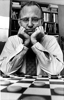 Marion Tinsley posing in a board of checkers with both of his hands in his chin and wearing a white long-sleeved polo and a black tie.