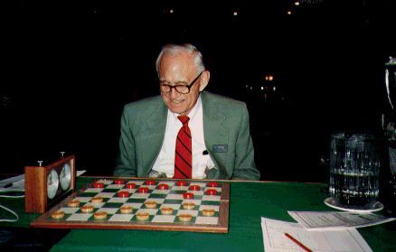Marion Tinsley happily playing checkers and wearing a green suit along with a white long-sleeved polo and a red tie.
