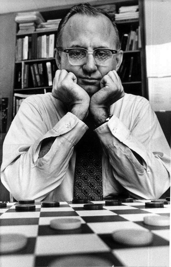 Marion Tinsley posing in a board of checkers with both of his hands in his chin and wearing a white long-sleeved polo and a black tie.