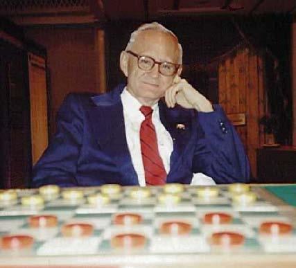 Marion Tinsley posing near a checkers board with his hand in his face and wearing a blue suit and a white long-sleeved polo with a red tie.