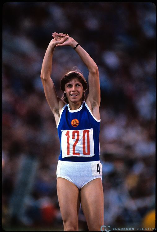 Marita Koch smiling while raising her hands and wearing a blue top and white shorts