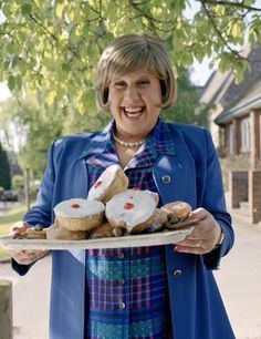 Marjorie Dawes with a big smile while carrying a plate with food, with short blonde hair, wearing a necklace, and a blue blazer over a multi-colored checkered polo shirt.