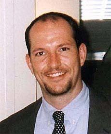 Mark Bingham smiling while wearing a black coat, blue long sleeves, and polka dot necktie