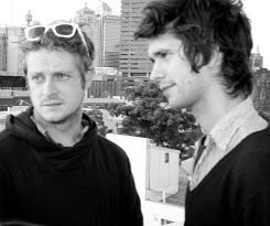 Mark Bradshaw and Ben Whishaw with serious faces while looking at something. Mark is wearing sunglasses on his head and a black jacket while Ben is wearing a black jacket over a white polo shirt.