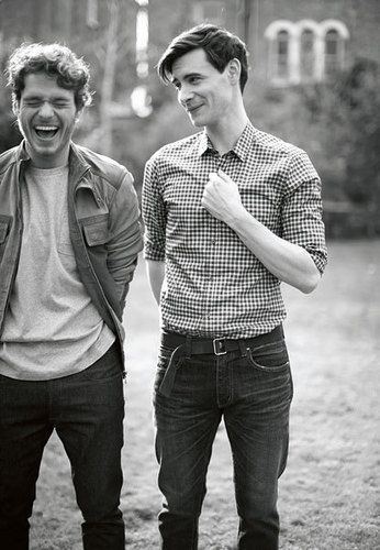 Mark Bradshaw is laughing while Ben Whishaw with a tight-lipped smile with his hand on his chest. Mark with curly hair, wearing a denim jacket over a gray shirt and black pants while Ben is wearing a folded checkered long sleeves, black belt, and black pants.