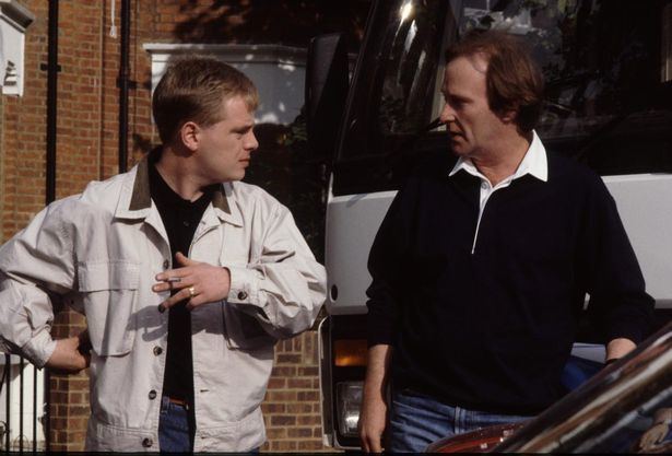 Mark Farmer smoking while wearing a white jacket and black t-shirt and Dennis Waterman wearing a black jacket