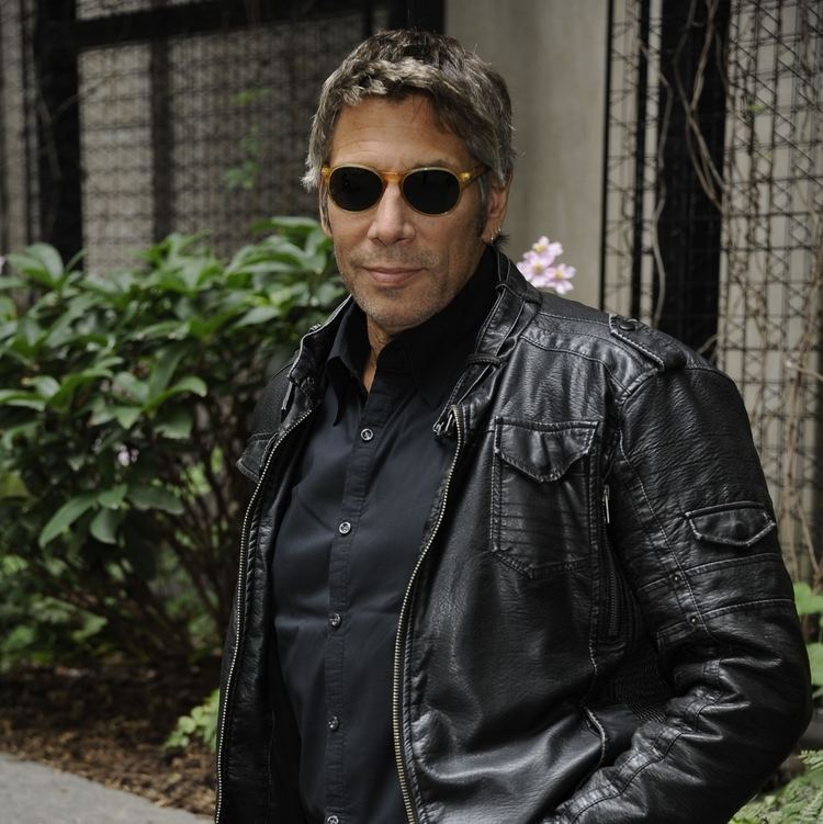 Mark Goodman with a tight-lipped smile while wearing a black leather jacket, black polo, and black sunglasses