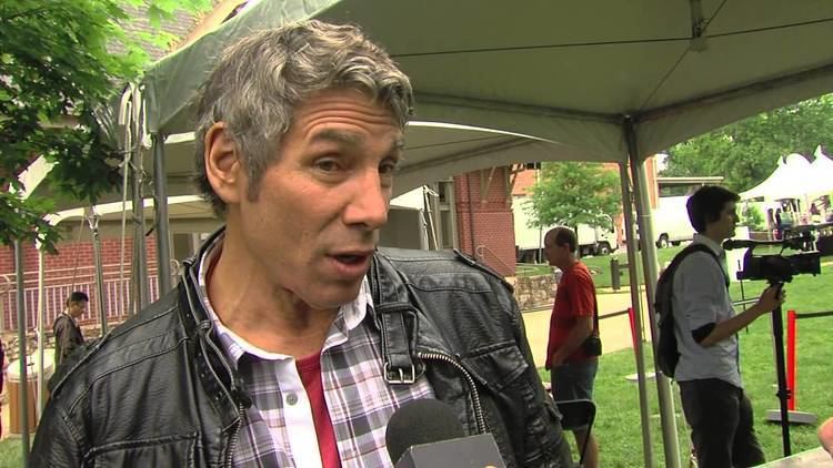 Mark Goodman, in one of his interviews, wearing a black leather jacket and a white, black, violet checkered polo