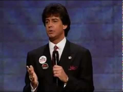 Mark R. Hughes talking on stage during a Herbalife presentation and wearing a black suit and tie.