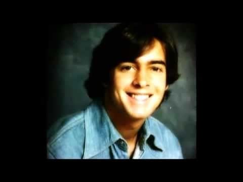 A younger Mark R. Hughes with long hair smiling and wearing a jean shirt.