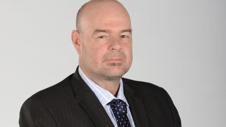 Mark Robinson's tight-lipped smile while wearing a black coat, light blue long sleeves, and polka dot necktie