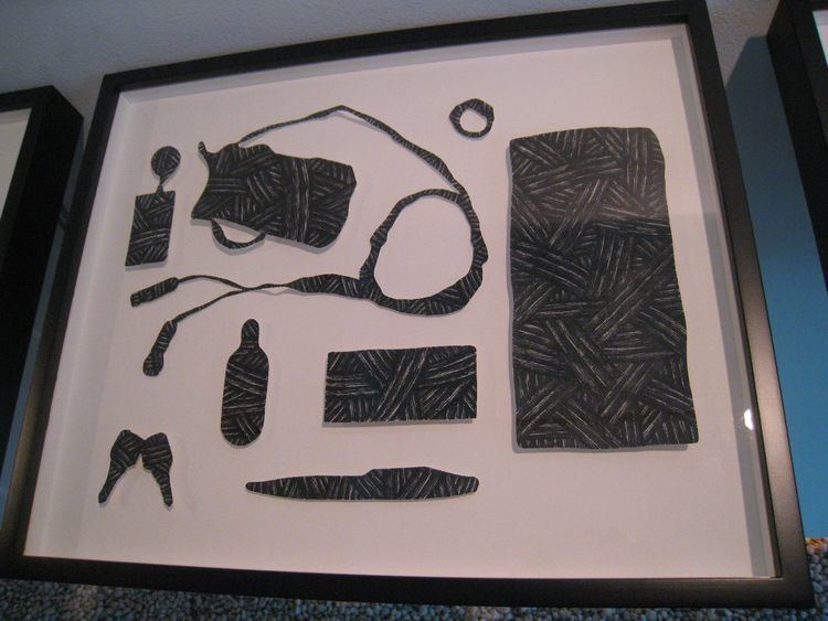 In a black frame on a wall is an art created by Mark Salvatus, it has black striped, shaped things, from left, a Bag, Pliers, Watch, Bracelet, Shoes, Hammer, leaf, an Id with key and lace framed in black.