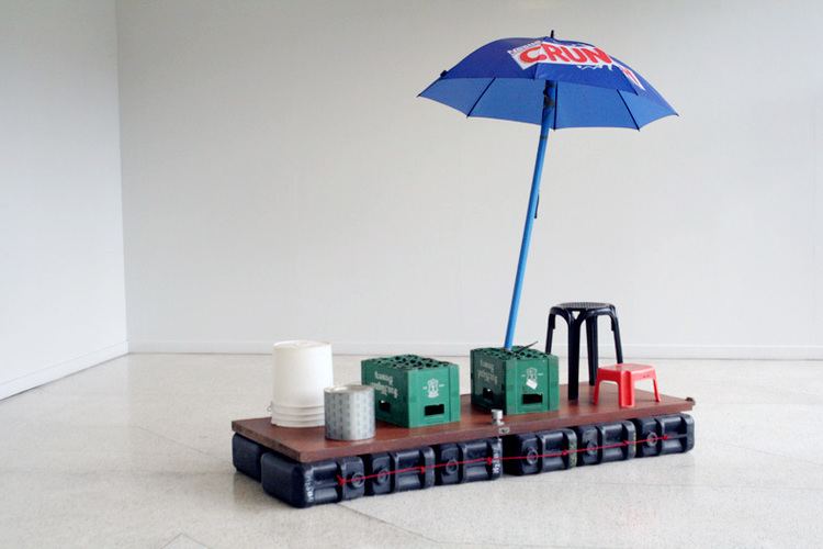 In a white room, is a brown miniature raft with black container at the bottom with red thread, red low chair and a tall black chair, with a white container, green beer cases and blue umbrella with crunch written on on.