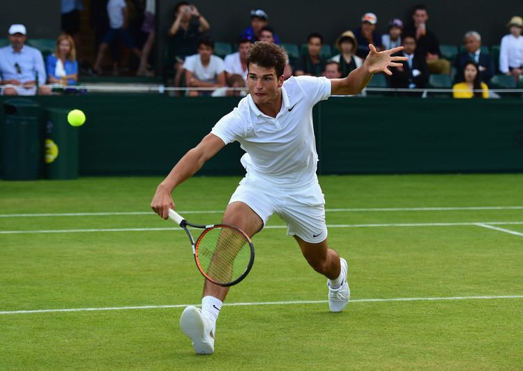 Marko Osmakcic playing tennis while wearing a white polo shirt, white shorts, and white rubber shoes