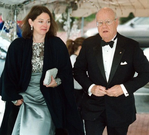 Deborah Adair Clarke with a serious face while holding a pouch bag while Forrest Mars Jr. with a serious face while fixing his black coat. Deborah is wearing earrings, a black coat, and a mint green dress while Forrest is wearing eyeglasses, a ring, a black coat over white long sleeves, a black bow tie, and black pants.
