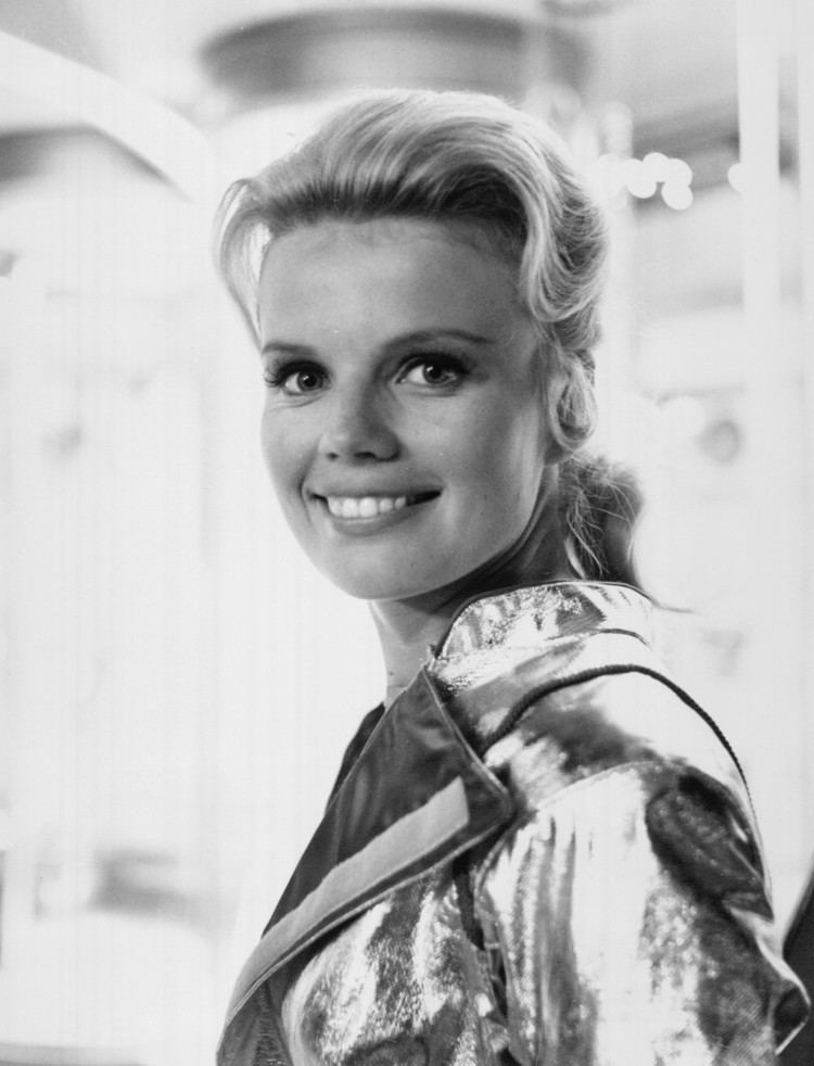 Marta Kristen with a smiling face and blonde hair while wearing a glossy top.
