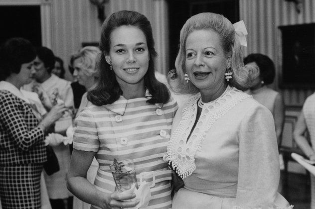 Martha Beall Mitchell (right) with Julie Nixon Eisenhower (left) are smiling together while their hands are around each other with a few other women in their background in an old photograph. Martha has white hair with a tie ribbon on it wearing dangle earrings, a pearl necklace, and a white vintage long sleeve dress while  Julie is holding a glass with tissue in her right hand, has black hair and wearing a ring in her right hand finger, a dangle earring and a striped vintage dress
