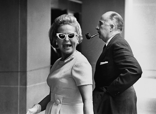 Martha Beall Mitchell (left) along with John Mitchell (right) are walking while Martha is talking to someone in an old photograph. Martha has a blonde hair wearing sunglasses, earrings, pearl necklace, white gloves and a white vintage dress while John has a smoking pipe on his mouth, wearing a white polo long sleeve under a black coat with a a white handkerchief in its pocket