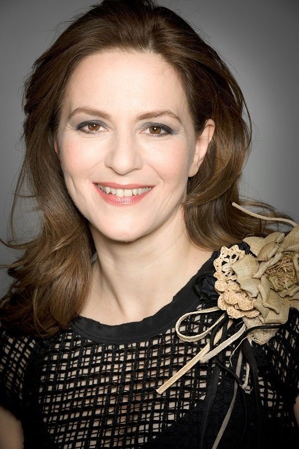 Martina Gedeck smiling while wearing a black blouse with a beige flower on the side