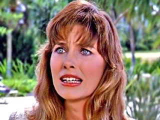 Mary Kate McGeehan with an angry look, brown wavy hair, gray eyes, and trees in the background while she is wearing a blouse