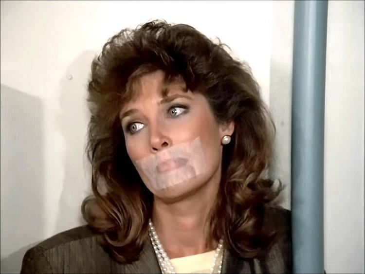 Mary Kate McGeehan in a scene from the 1982 tv series, Knight Rider. Mary Kate is looking on the left side with a sad face, wavy hair, and tape on her mouth while wearing a pearl necklace and yellow blouse under a dark brown coat