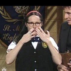 Mary Katherine Gallagher (left) is serious, has black hair, both hands on her mouth, a ring on her right hand, wearing eyeglasses, a white school uniform under a black vest with the school patch, a dark-blue bow tie, and a red headband. Gabriel Byrne (right) is serious while looking at Mary Katherine Gallagher, he has black hair, both hands holding a clipboard, wearing black long sleeves under a black suit.