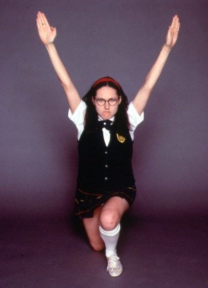 Mary Katherine Gallagher is serious, both hands up, right knee bent, she is wearing an eyeglass, white school uniform under a black vest with the school patch, a dark-blue bow tie, a red headband, a plaid skirt, white knee socks, and white shoes.