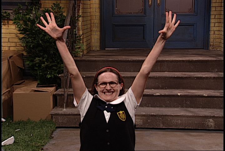 Mary Katherine Gallagher is smiling, has black hair, behind her is a blue door, stairs, and boxes, both hands up, ring on her right hand, she is wearing an eyeglass, white school uniform under a black vest with the school patch, a dark-blue bow tie, and a red headband.