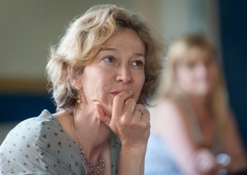 Matilda Ziegler looking at something while hand on her chin and wearing light blue and black blouse, necklace, earrings, and ring