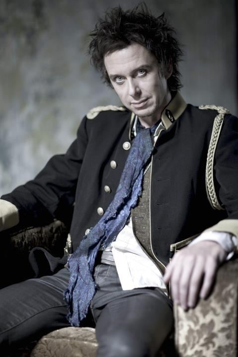 Matt King with a serious face while sitting on a couch, with messy hair, wearing a black coat with a blue scarf, and black pants.