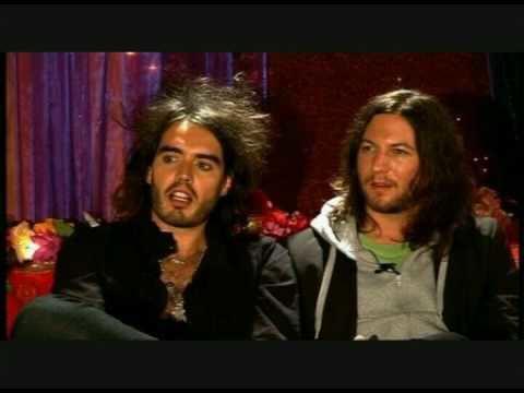 Matt Morgan sitting on the couch with Russell Brand while wearing a black coat, gray jacket, and green t-shirt