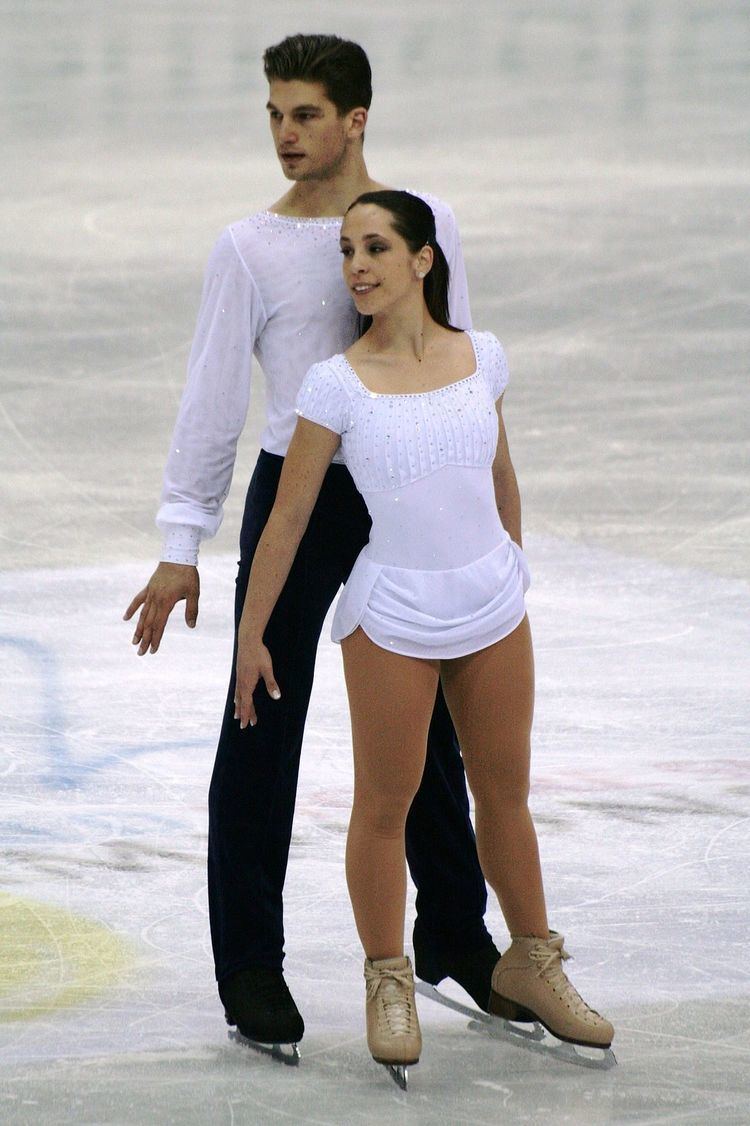 Nicole Della Monica and Matteo Guarise are dancing on the ice while looking at the left side during the 2012 World Championships. Nicole is wearing a white dress and beige skating shoes while Matteo is wearing a white long sleeve shirt, black pants, and black skating shoes